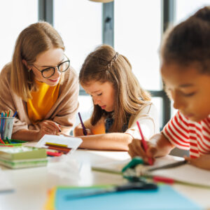 Smart woman teacher in  glasses helping students to to schoolwork during lesson in light classroom at school